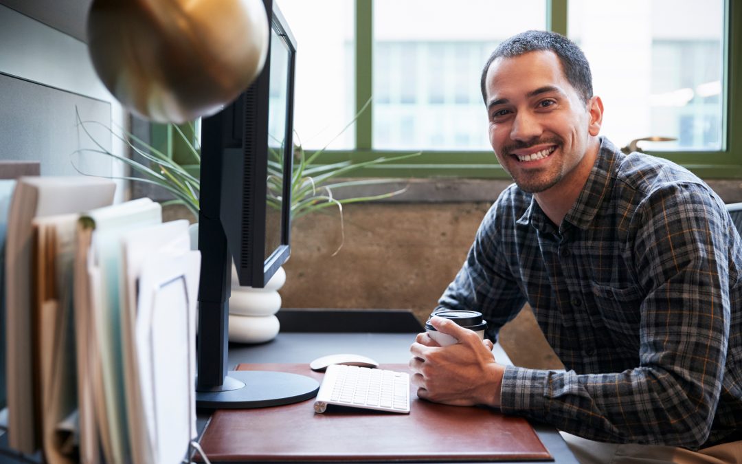 A smiling person in a plaid shirt sits at their desk, engaging with everpayer's financial solution, with a keyboard and coffee cup beside their monitor.