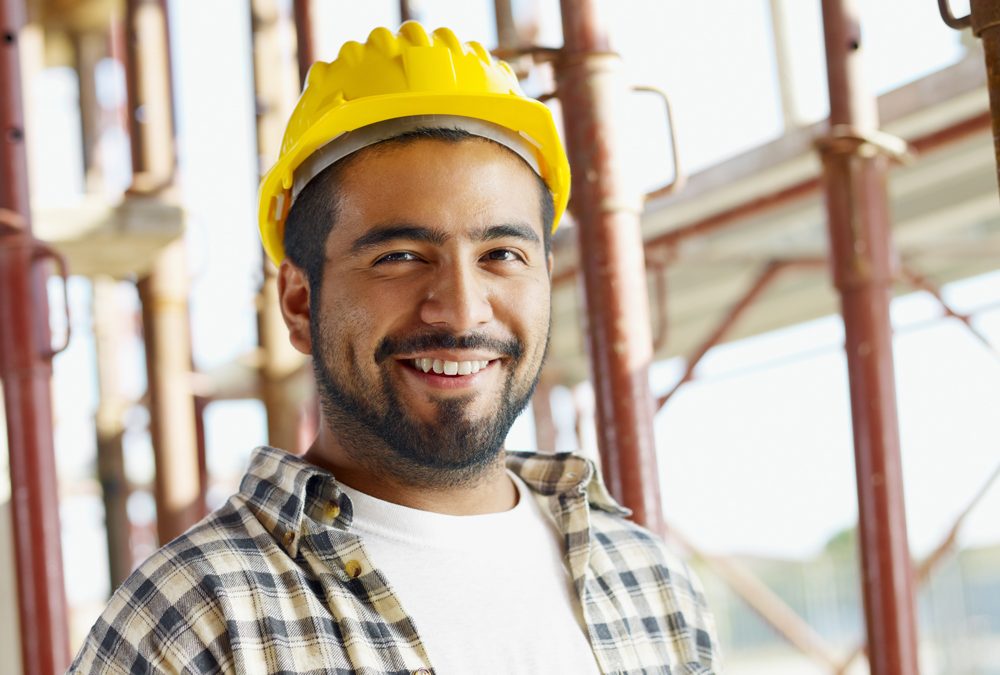 Smiling construction worker in a yellow hard hat and plaid shirt at a building site with scaffolding in the background, pondering efficient money transfer solutions with Everpayer to help streamline project finances.