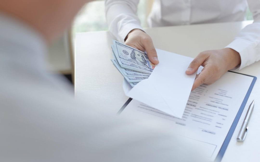 Person handing an envelope filled with cash over a document on a clipboard to another person.