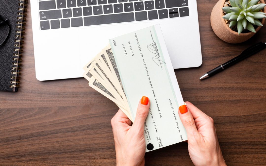 Hands holding cash and a check above a wooden desk with a laptop, notebook, pen, and a small potted plant.