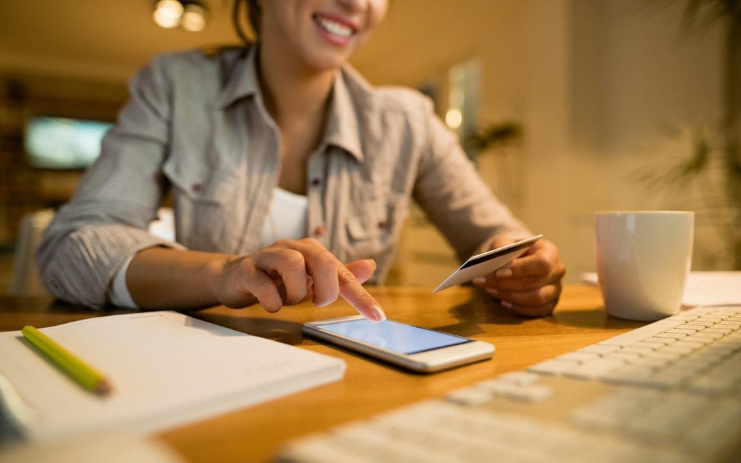 Smiling woman uses smartphone and holds a credit card, sitting at a desk with a keyboard and notebook.