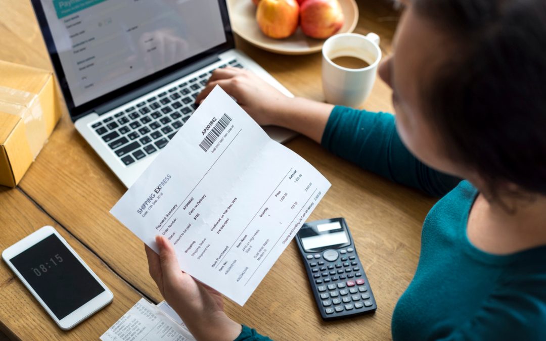 Person holding a shipping invoice at a desk with a laptop, calculator, phone, coffee, and apples.